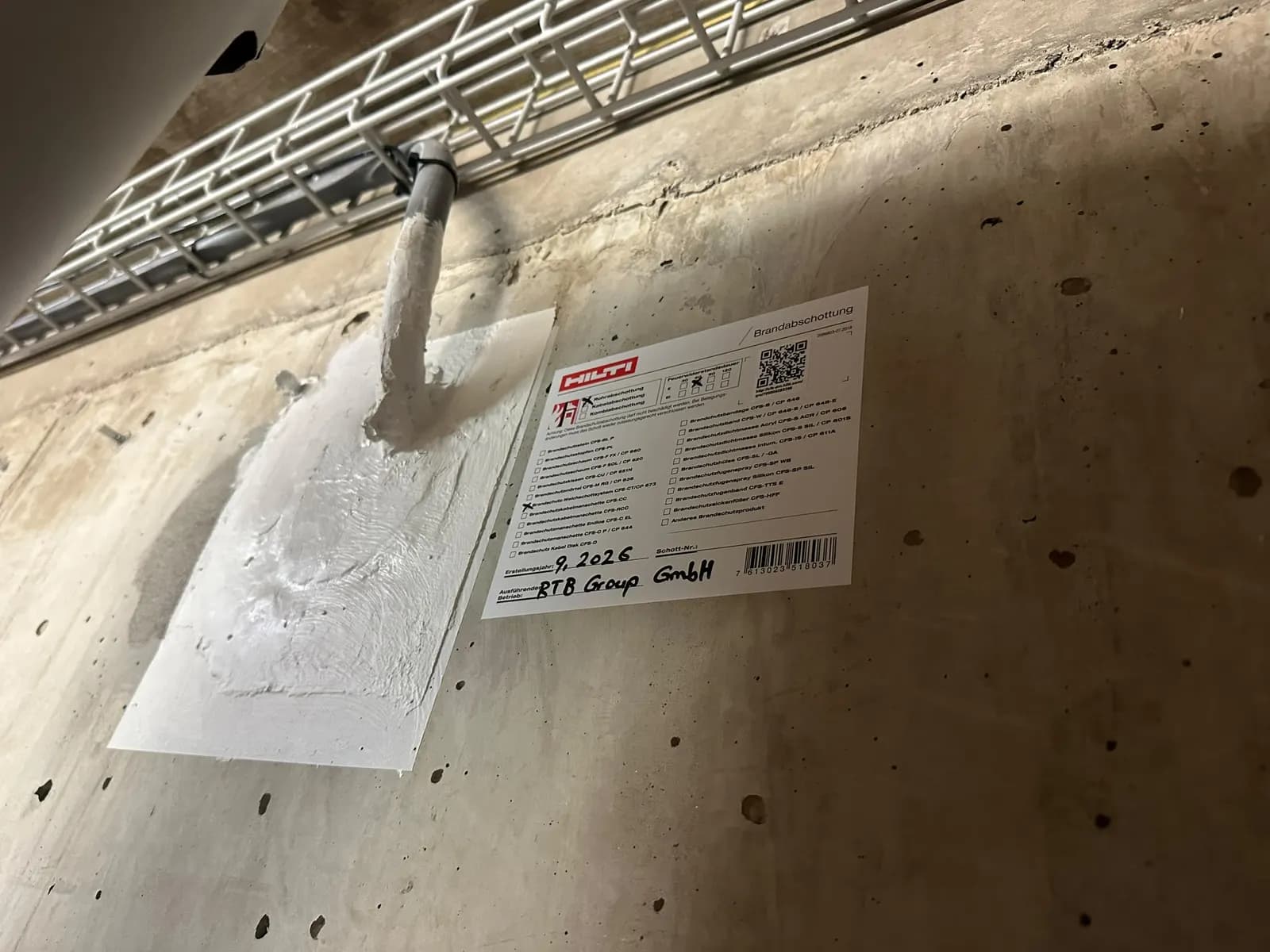Rohrabschottung mit Brandschutzkabelmanschette an einer Betonwand unter einer Kabeltrasse, Schottschild mit Erstellungsjahr und ausführendem Betrieb